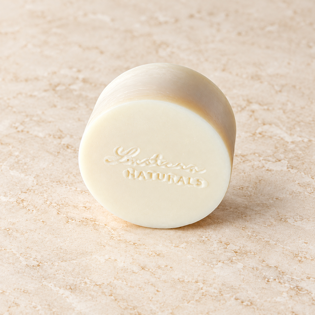 Angled view of one round ivory Aloe Quench conditioner bar with the Lustera Naturals stamp on warm ivory stone