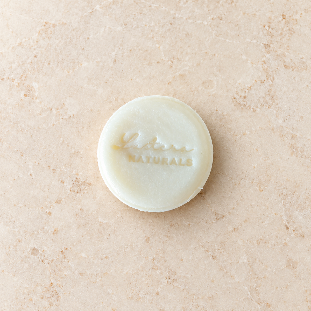 Top view of one round ivory Aloe Quench shampoo bar with the Lustera Naturals stamp on warm ivory stone